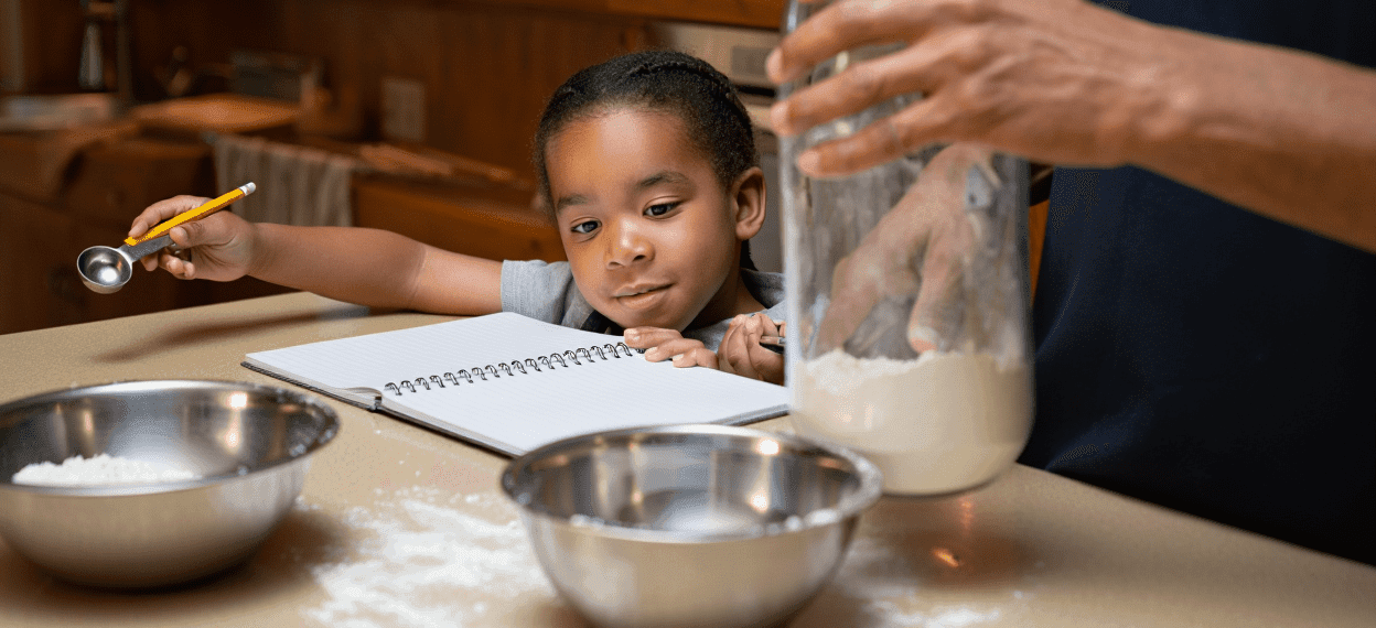 Child with measuring spoon, adult with flour, bowls, and notebook on counter.