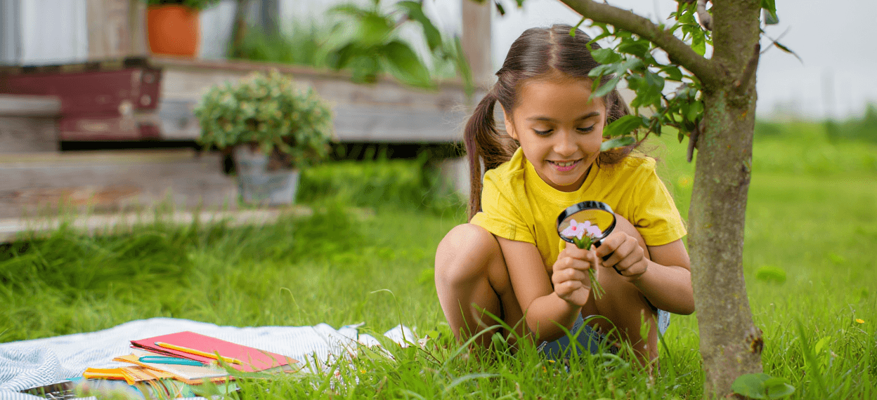 Child with pigtails examining pink flowers with a magnifying glass in grass.